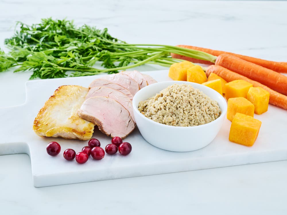 Wild Alaska pollock and pork minced cat food in a white bowl, styled on a white cutting board with sliced pork, a cooked fish fillet, carrots, leafy greens, cubed squash, and cranberries.