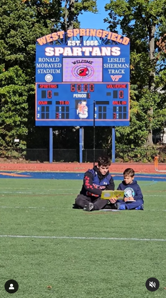 A high schooler and elementary school kid sit on a football field