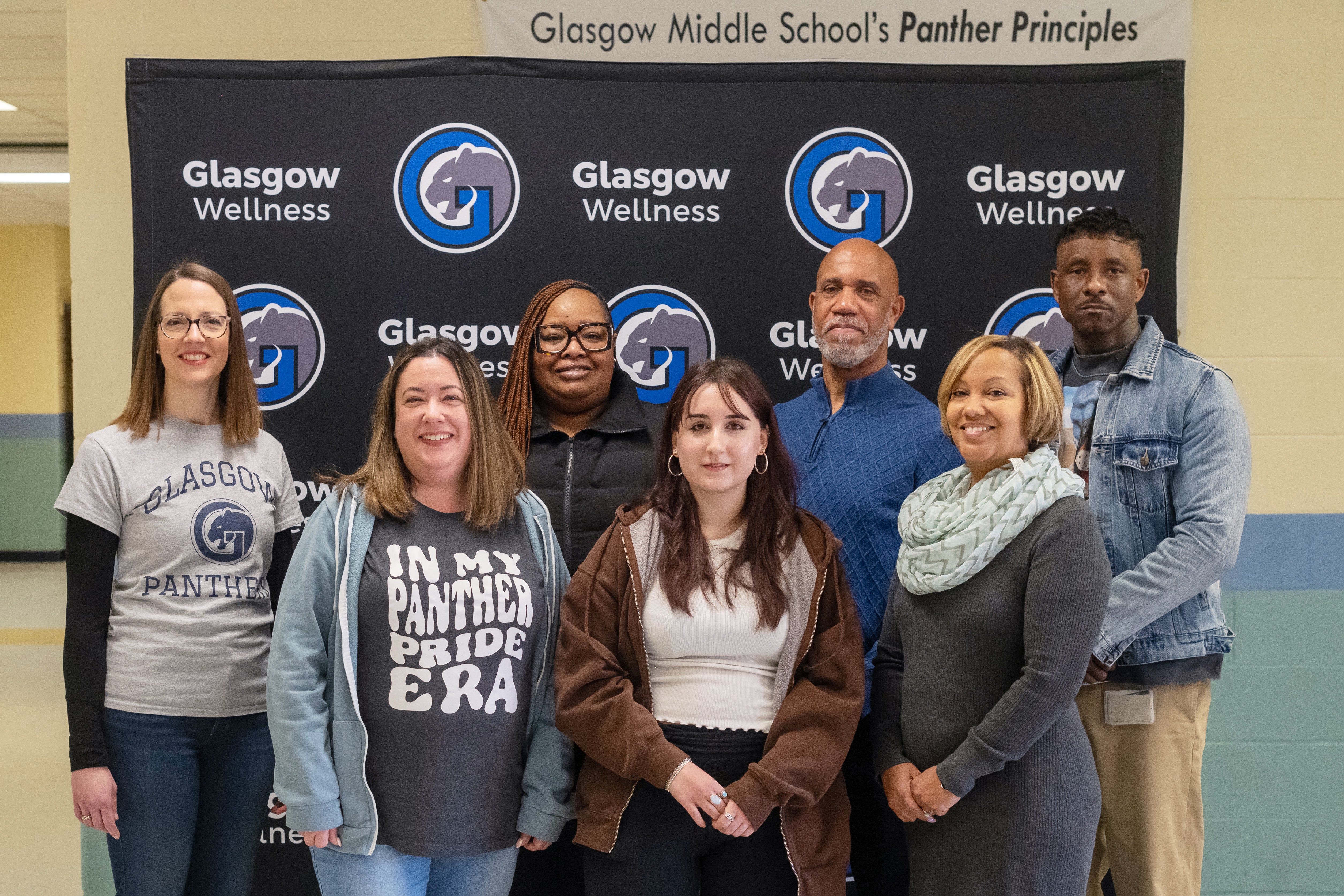 Seven adults smile in a group photo in front of a Glasgow Middle School backdrop.