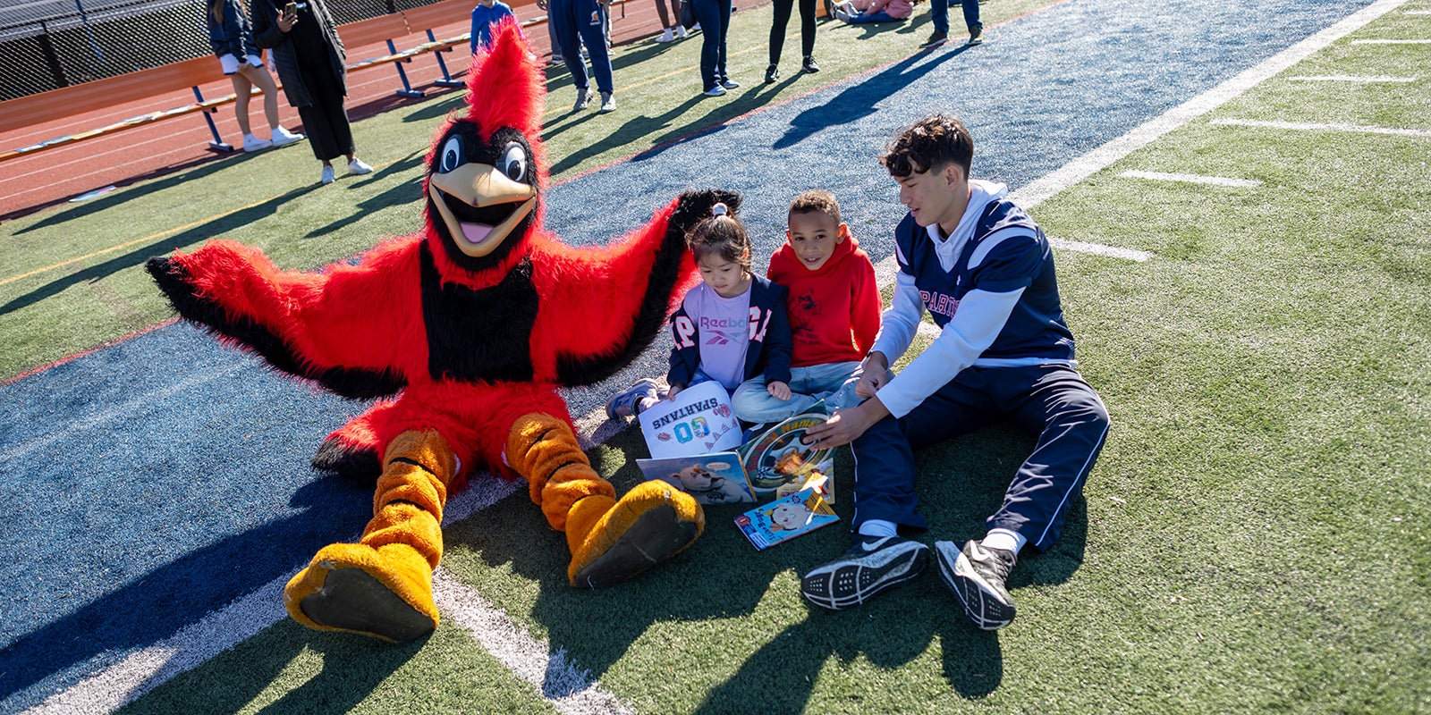 A cardinal mascot sits with two young kids and a high school football player on a field