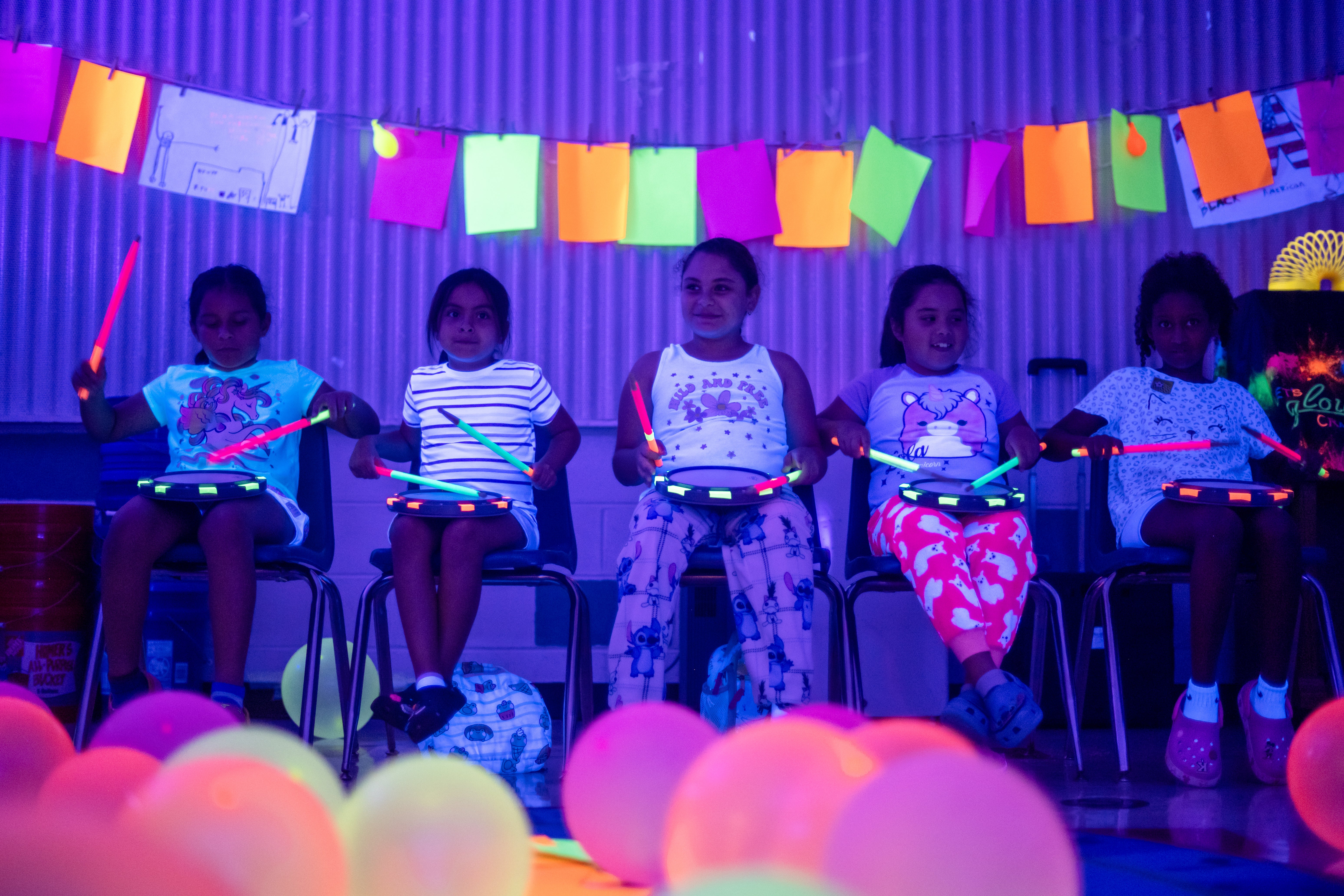 Students play drums under a black light, neon colors glow. 