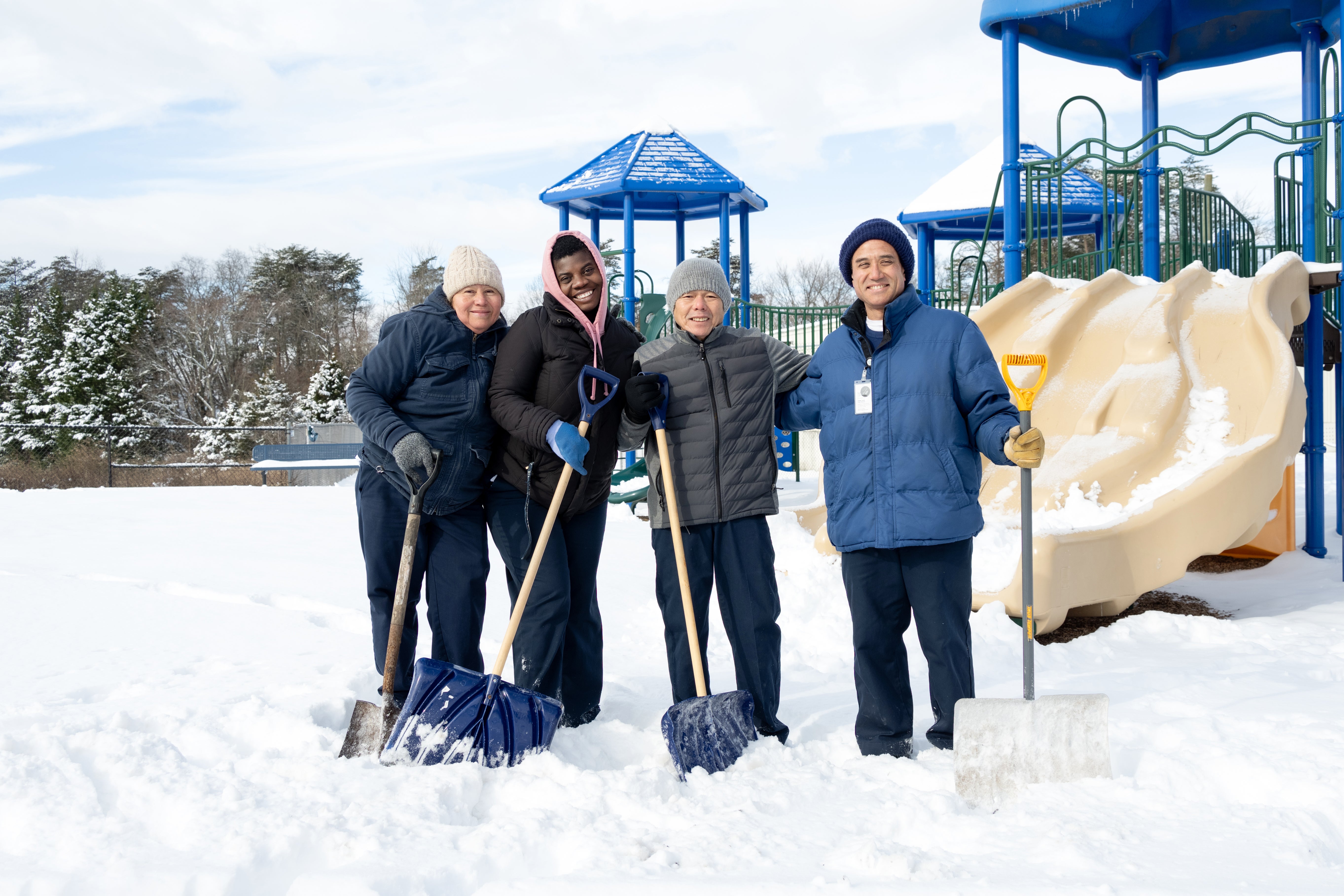 Custodians pose on a snowy playground with shovels. 