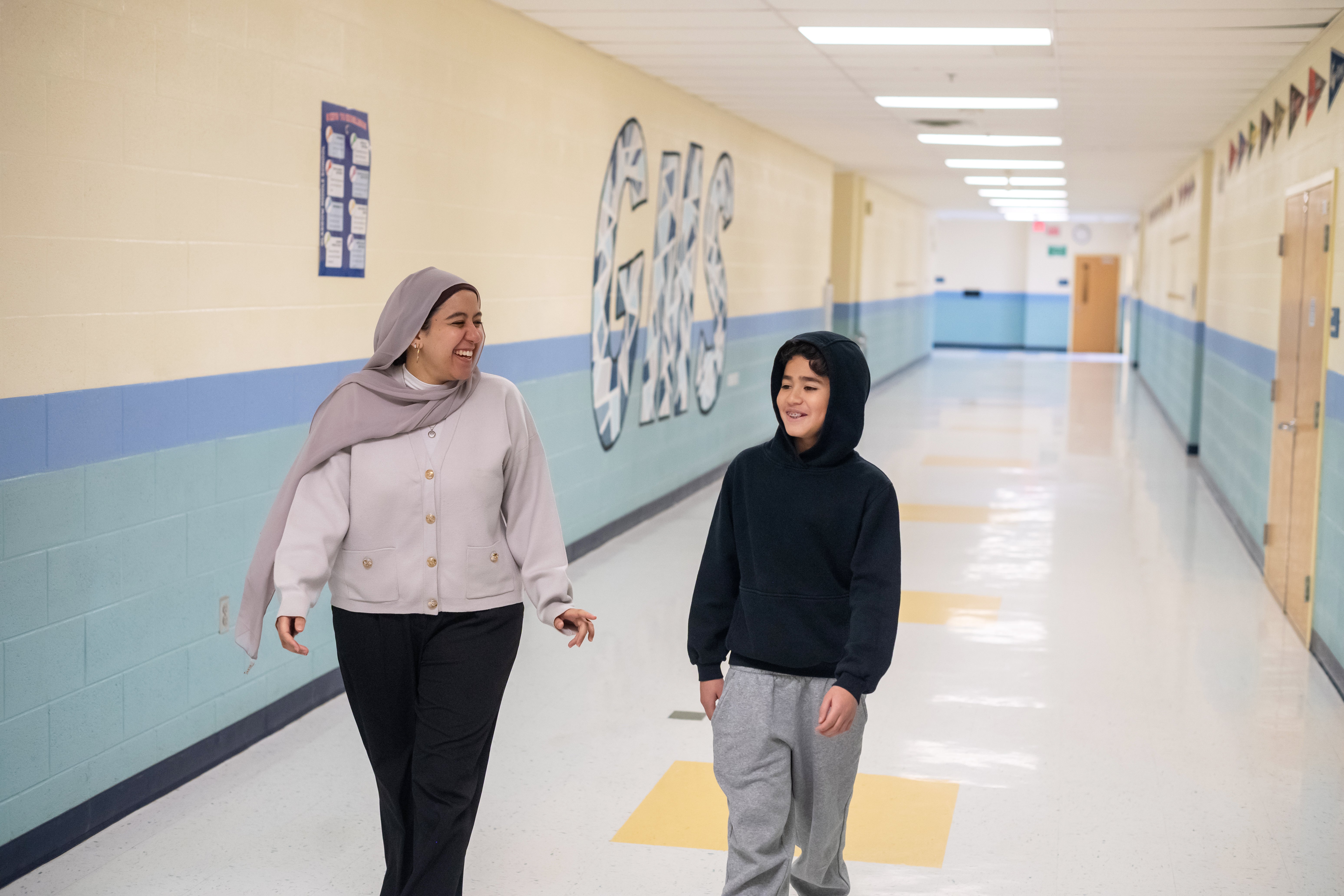 A young woman, left, laugh as she looks toward a grinning boy, right. The two are walking down a hallway.