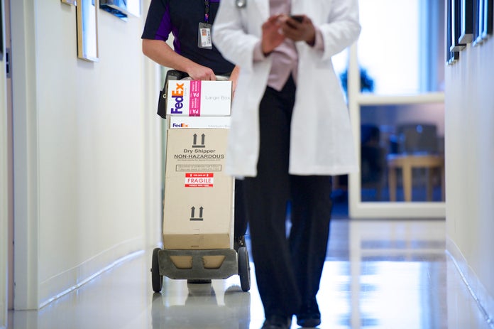 Doctor uses mobile phone while courier pushes stack of boxes in hallway