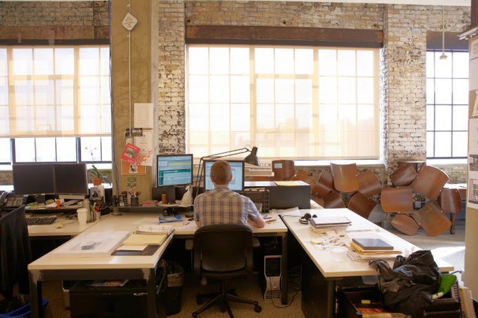 A man sits in a high-ceilinged brick and wood office facing his desktop computer
