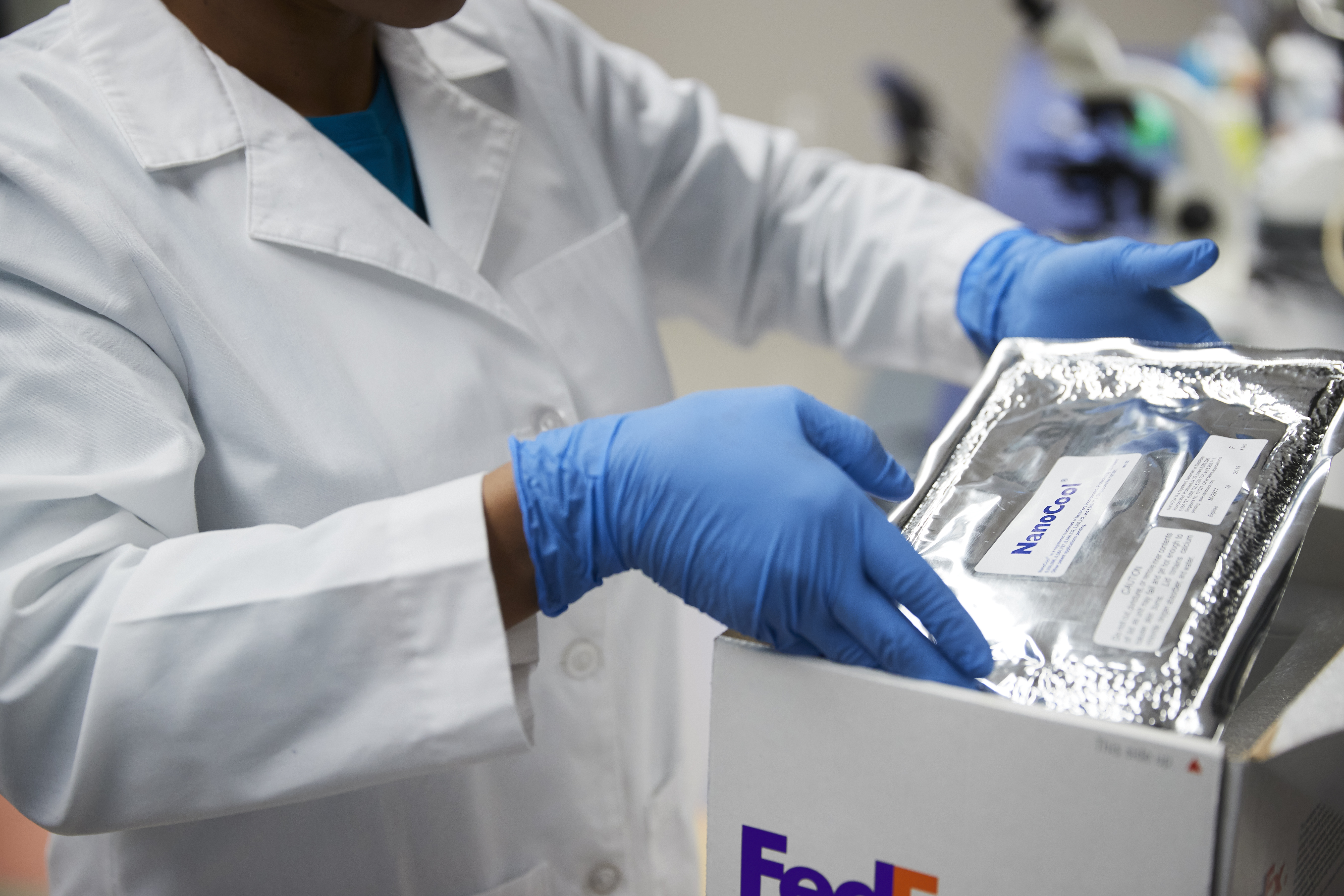 Person in a lab, wearing a white lab coat and blue gloves packing a cold item in a FedEx cold package.