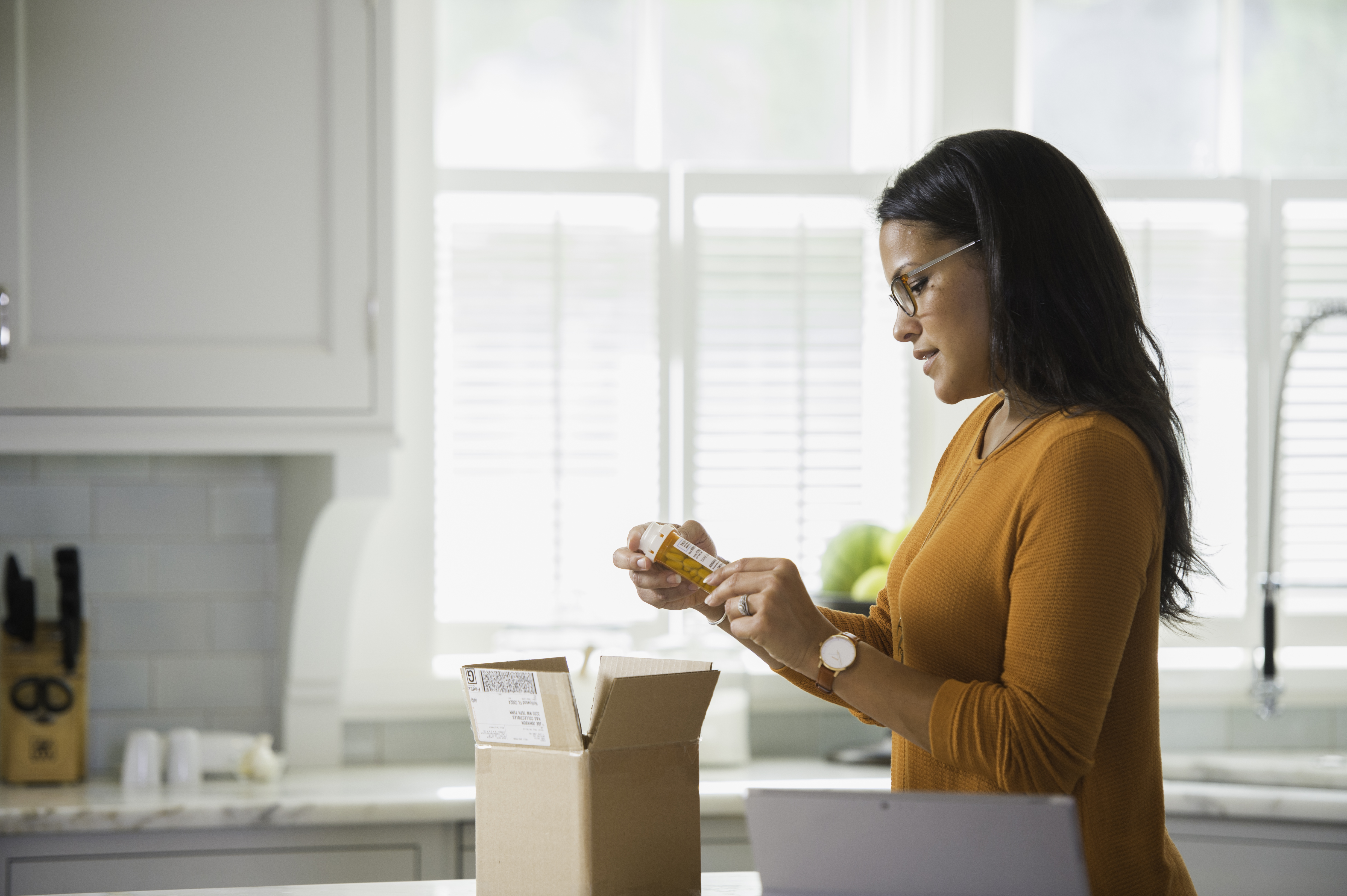 Woman looks at prescription bottle near package in kitchen