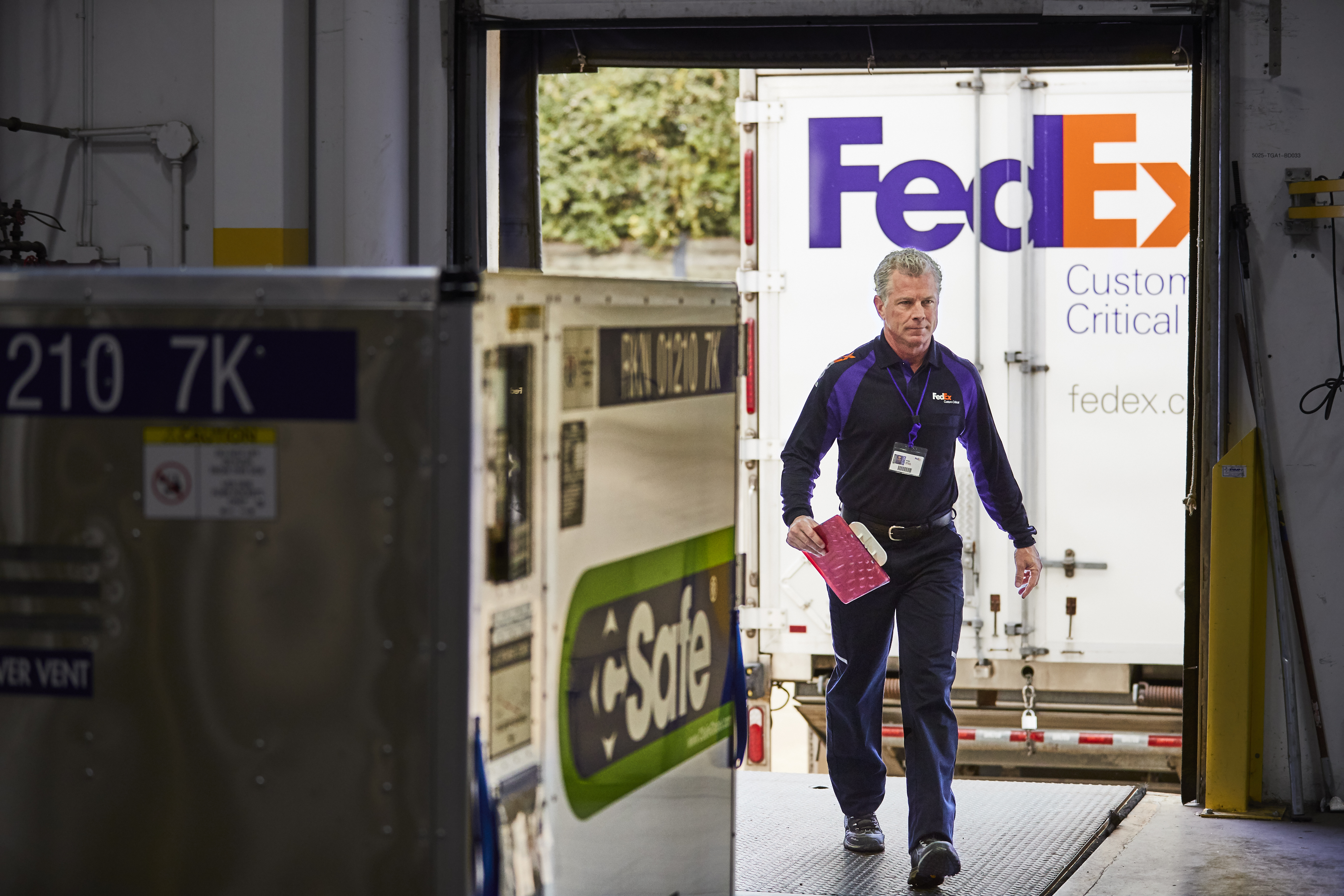 Estafeta da FedEx a afastar-se de um camião da FedEx em direção a um armazém e a um contentor C-Safe