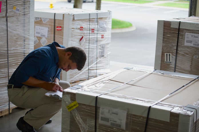 A warehouse worker checking cargo palettes completes paperwork on a clipboard.