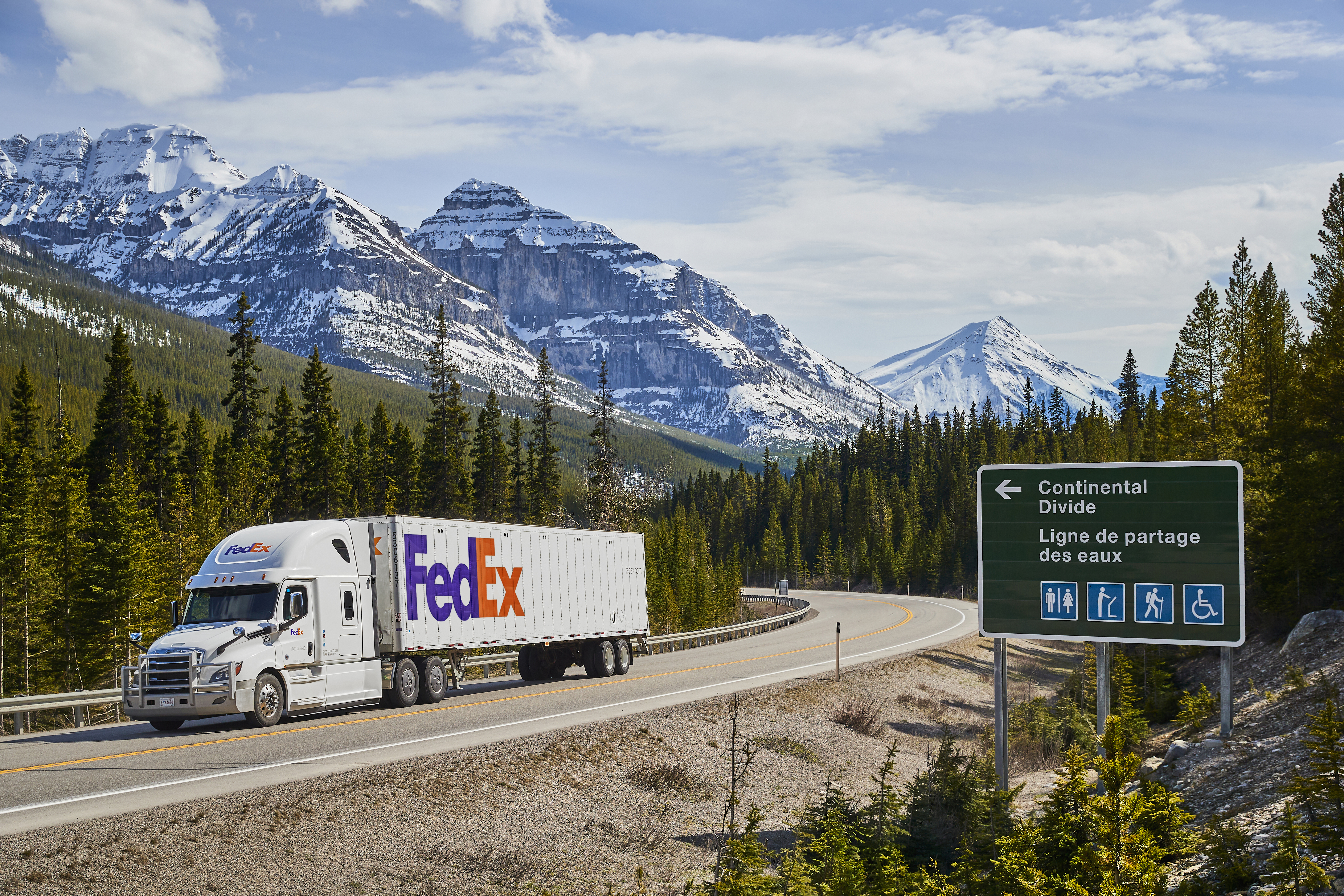 FedEx semi-trailer truck driving along a winding mountain road with evergreen trees and snowy peaks.