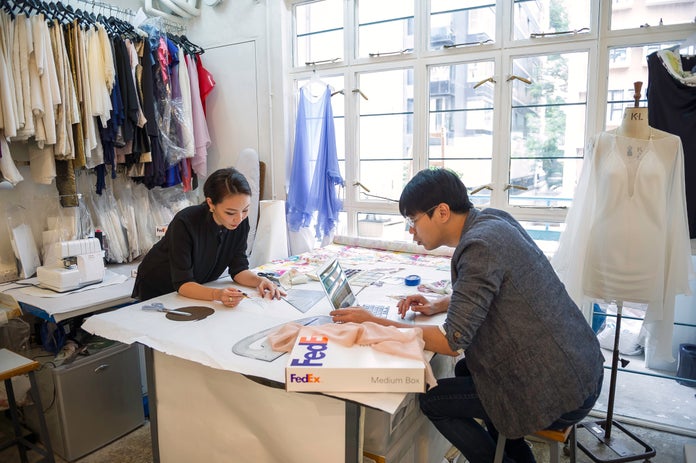 A clothing designer works with a pattern while her colleague uses his laptop.