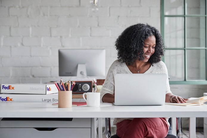 Woman using laptop with FedEx boxes on table.
