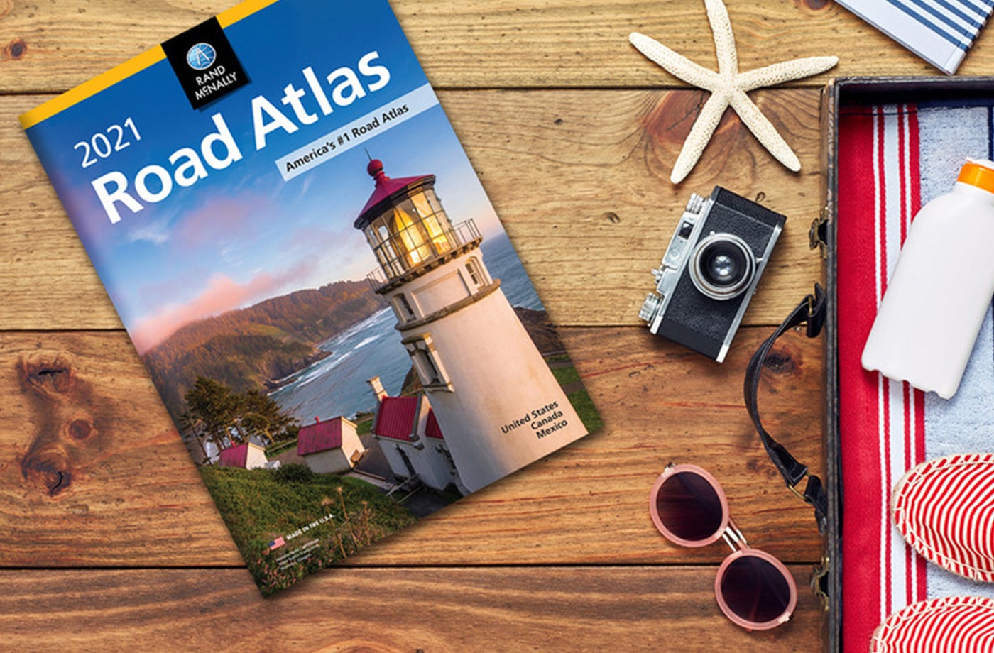 Rand McNally Road Atlas on top of wooden table along with sunglasses, a camera, sunscreen and a starfish