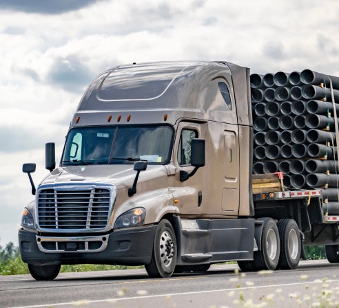 Semi-truck transporting large metal pipes on a flatbed trailer.