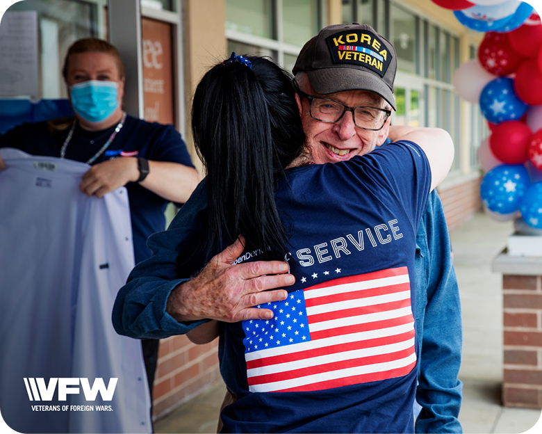 Older man wearing a Korea veteran hat hugging a woman in a U.S. flag shirt at a Veterans of Foreign Wars event.