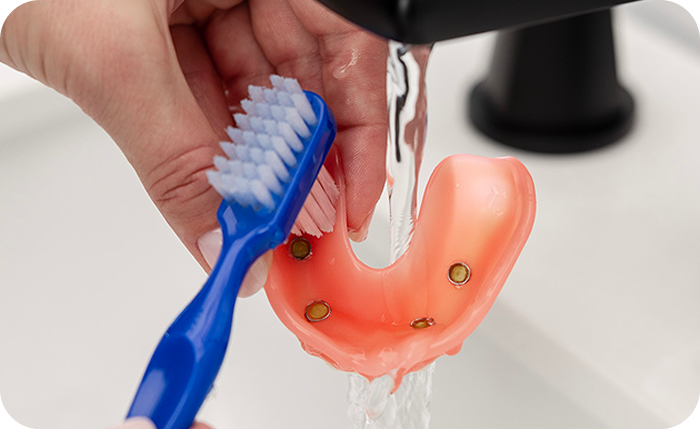 Alt text:
Person cleaning an implant-supported denture with a toothbrush under running water.