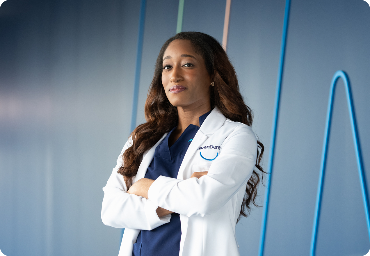 Aspen Dental professional wearing a white coat and navy scrubs standing with arms crossed in a dental office.