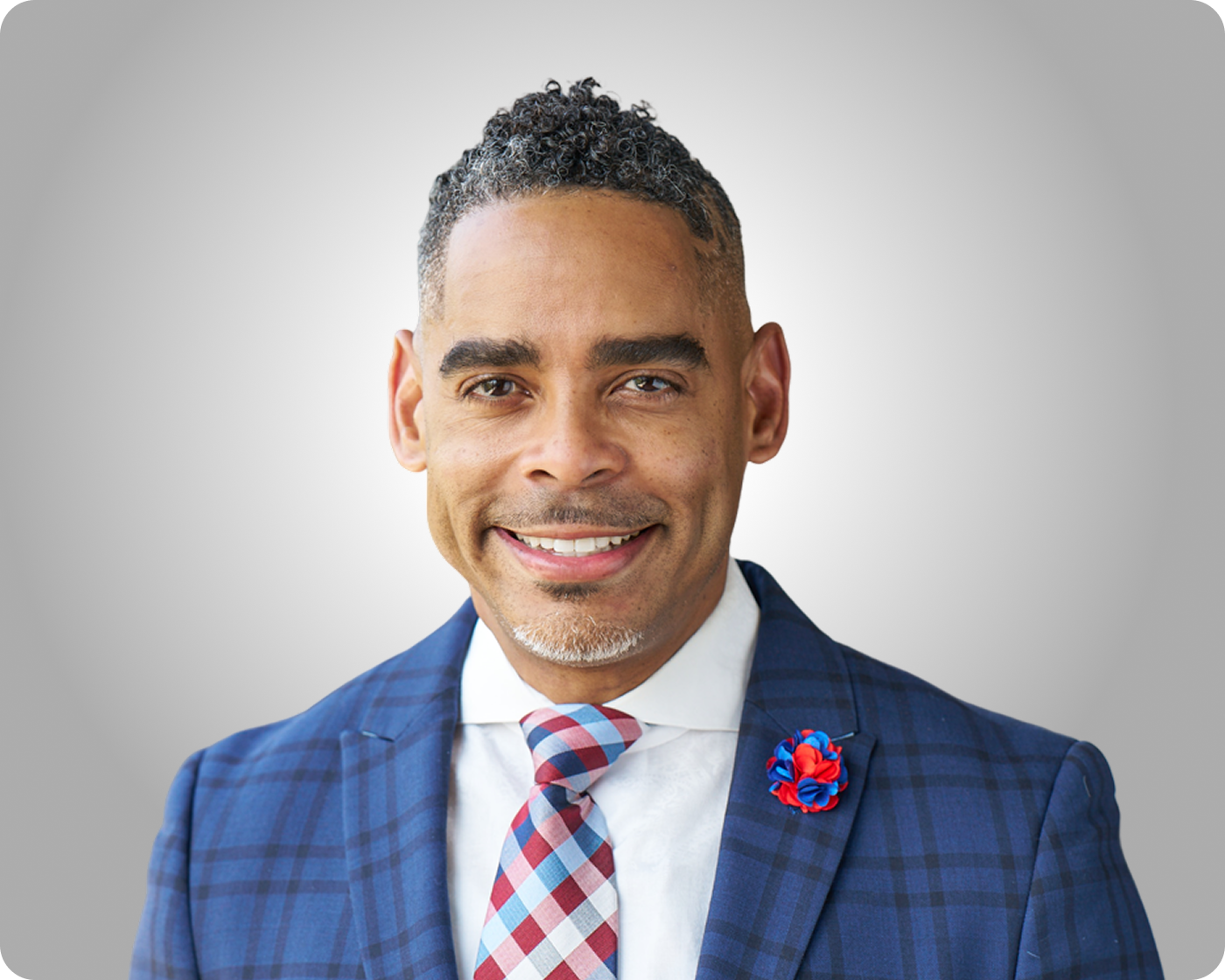 Professional headshot of a smiling man wearing a blue plaid suit and patterned tie against a gray background