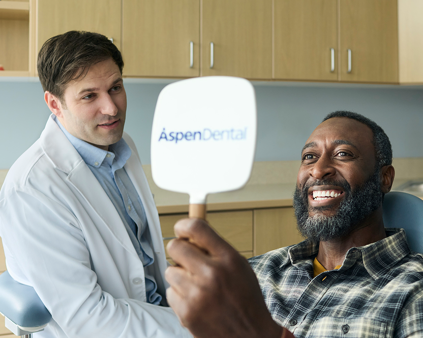 Alt text:
Smiling adult man holding an Aspen Dental mirror and viewing his smile during a dental visit.