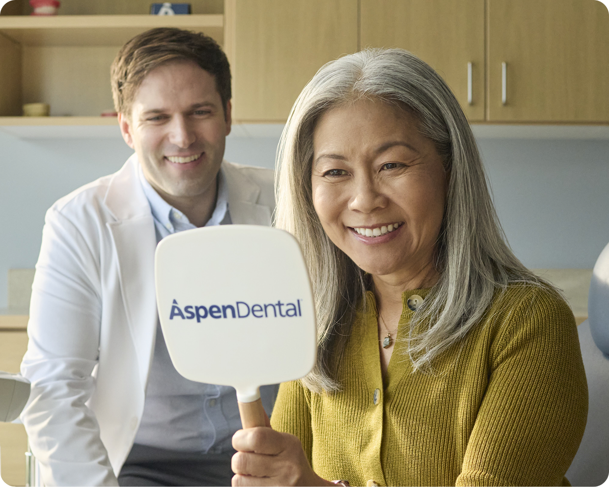 A smiling Aspen Dental patient is holding a hand mirror with the Aspen Dental logo while a dental provider sits beside her in a bright dental office.