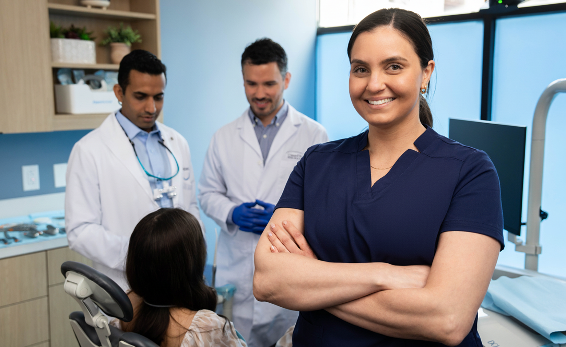 Alt text:
Aspen Dental clinician smiling with arms crossed in a treatment room while dental providers assist a patient in the background.