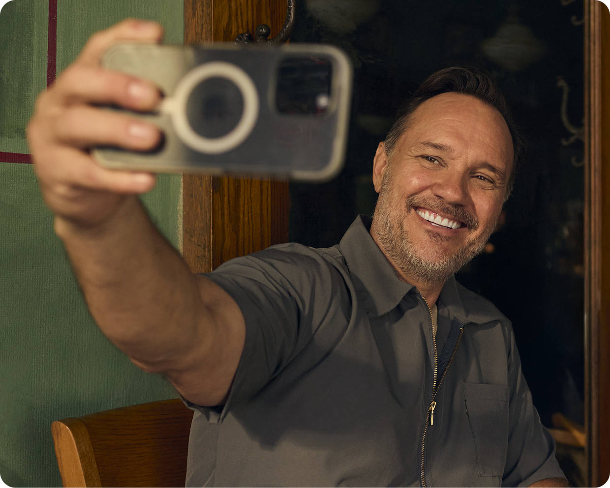 A smiling man is seated taking a selfie with his smartphone while indoors.