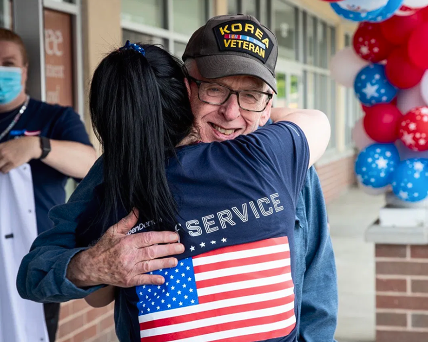 Alt text:
Older man wearing a Korea veteran hat hugging a volunteer wearing a U.S. flag “Day of Service” shirt.