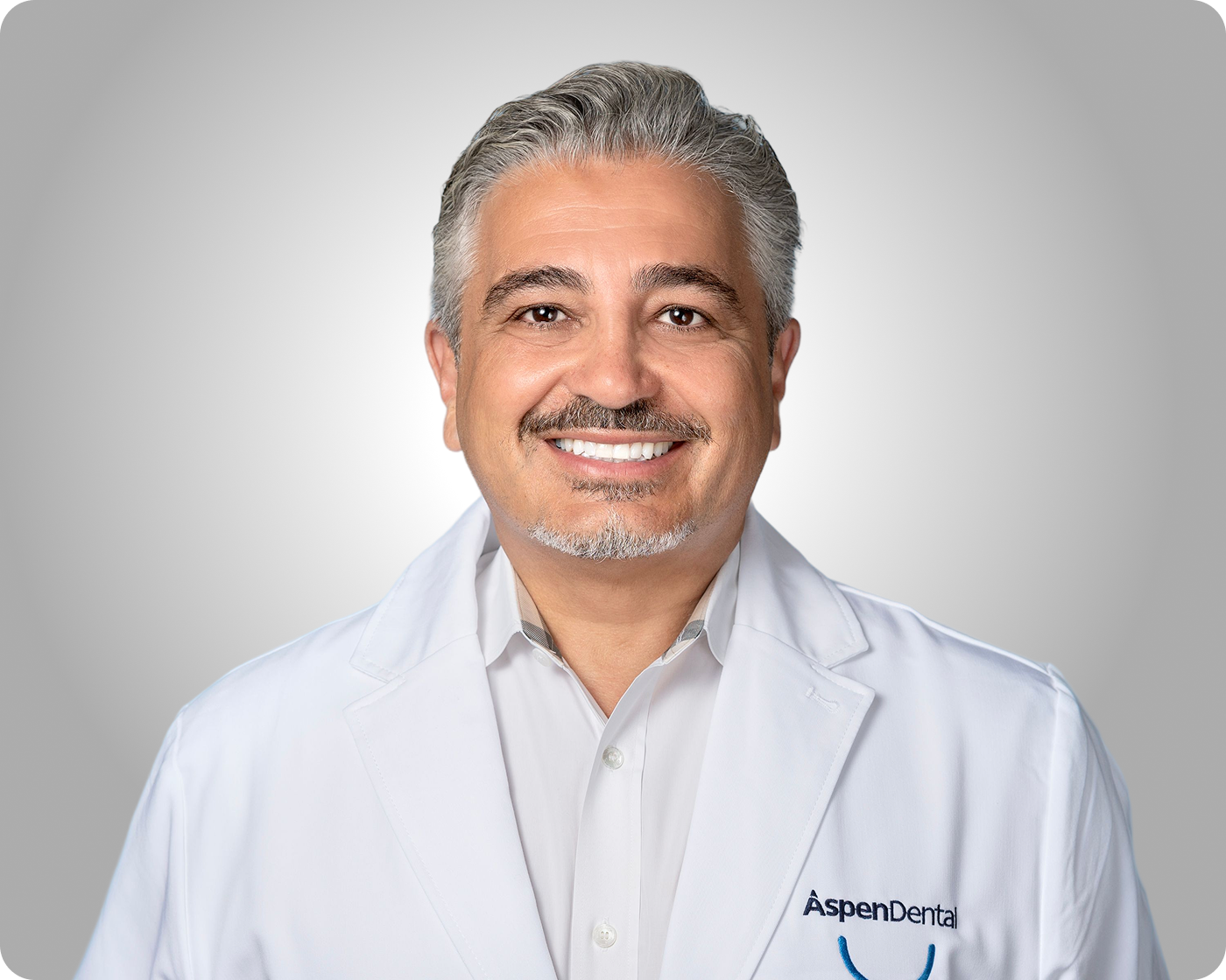 Professional headshot of a smiling male dental provider wearing a white coat against a gray background