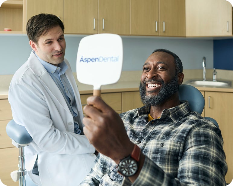 Patient holding an Aspen Dental mirror and smiling during a dental visit with a dentist nearby.