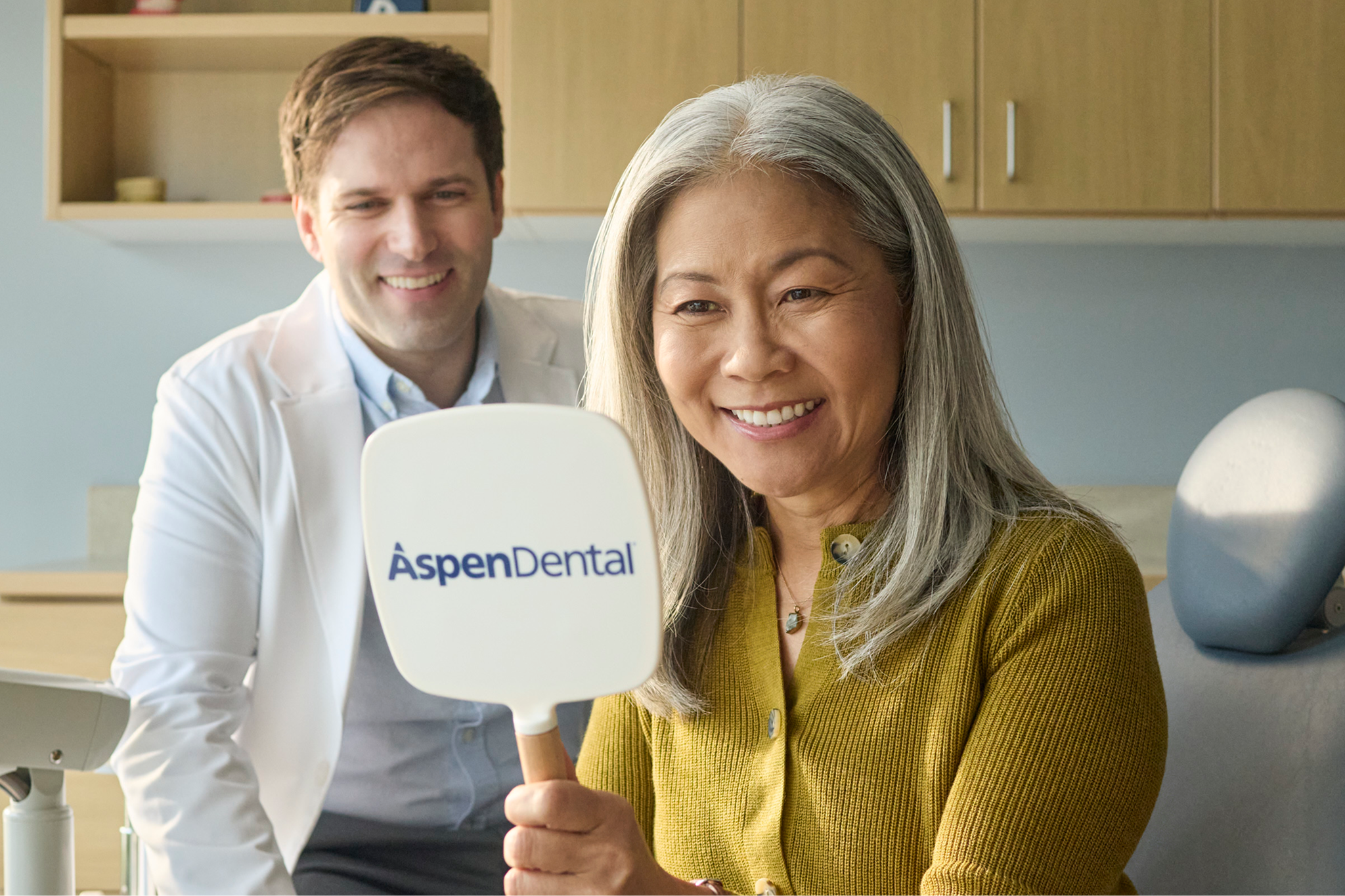 Alt text:
Patient smiling while holding an Aspen Dental mirror during a dental visit.