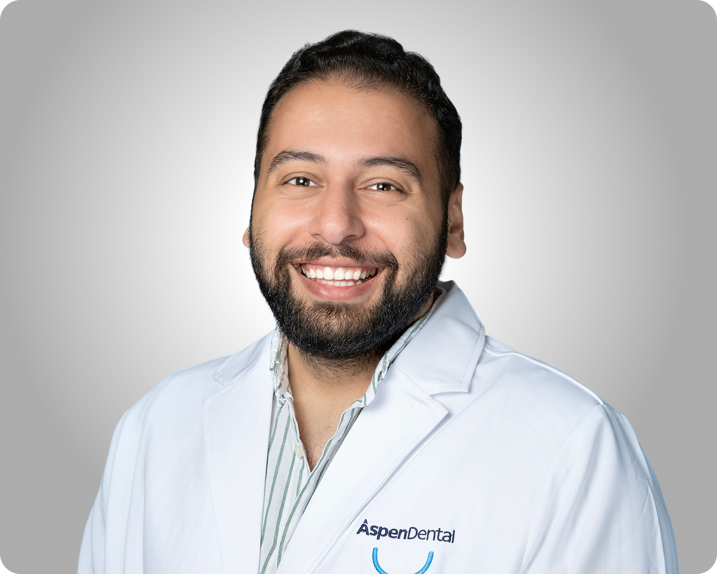Professional headshot of a smiling male dental provider wearing a white coat against a gray background