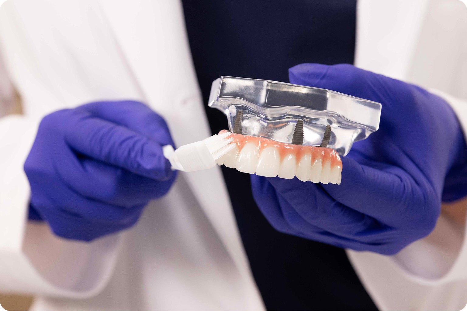 Gloved hands demonstrate proper cleaning of a dental implant model using a soft-bristled toothbrush.