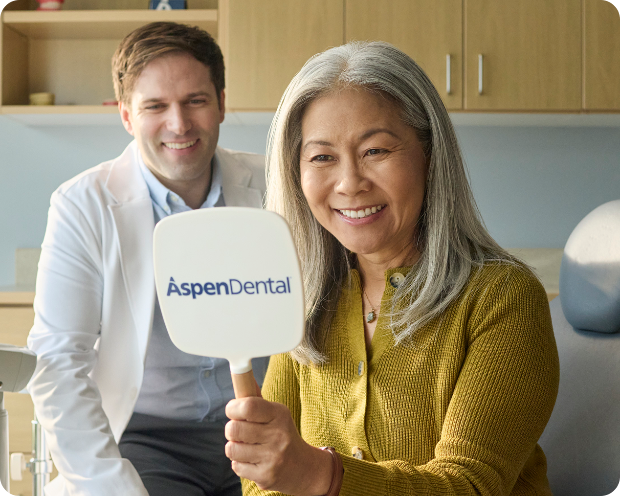 A happy patient looking into an Aspen Dental hand mirror to admire her restored smile while her dentist smiles in the background.