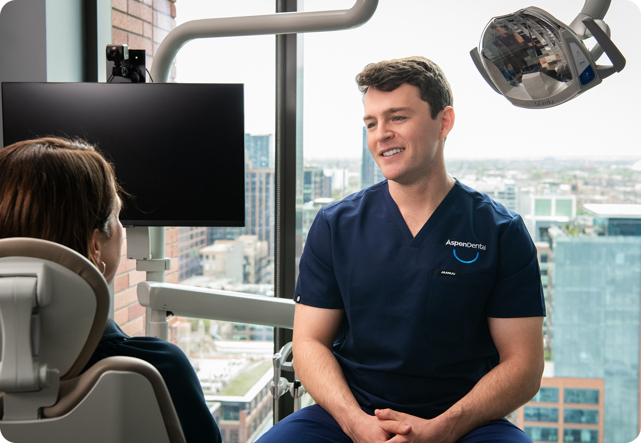 Smiling Aspen Dental professional in navy scrubs speaking with a patient seated in a dental chair.