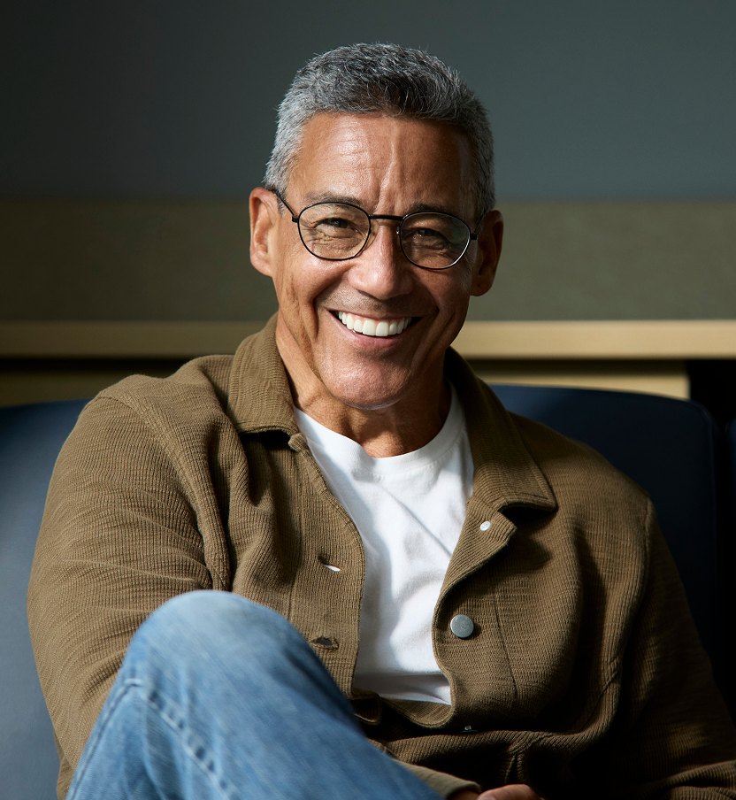 Alt text: Smiling adult man wearing glasses and a casual jacket seated indoors with a relaxed, confident expression.