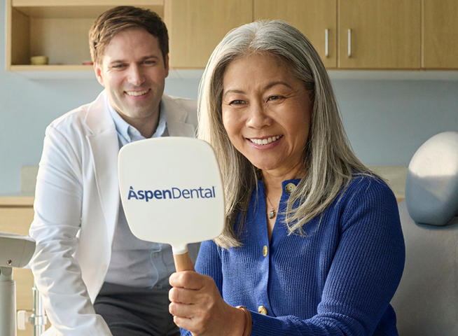 A happy aspen dental patient admires her smile in a mirror with her dentist