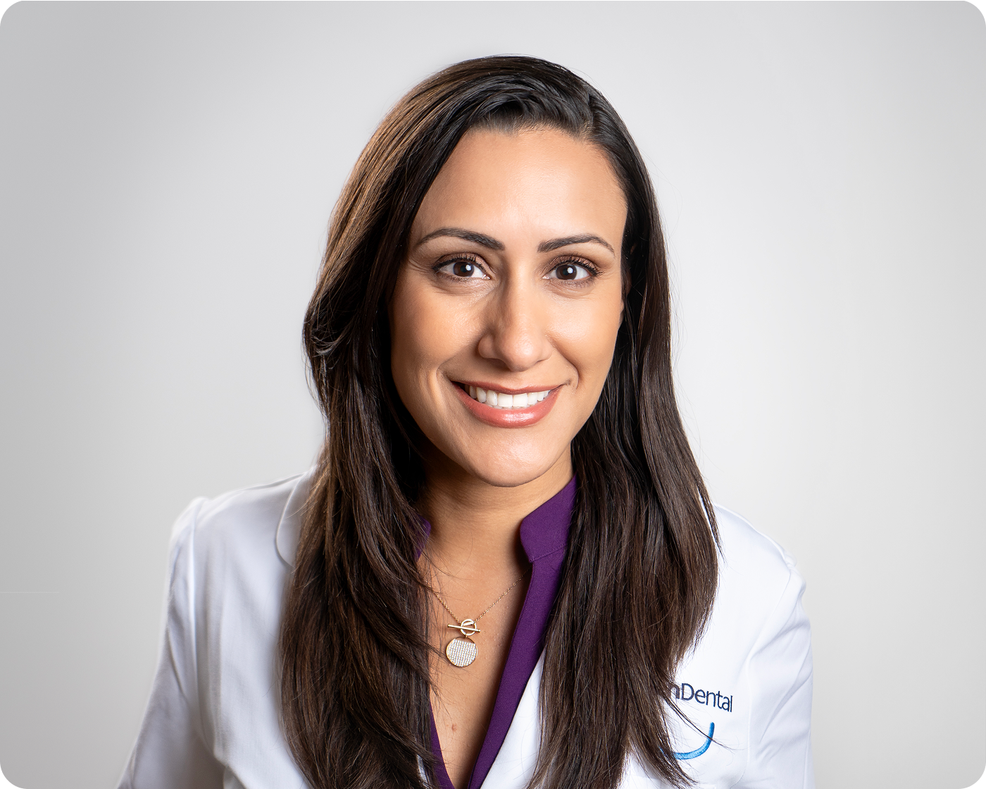 Professional headshot of a smiling female dental provider wearing a white coat against a gray background