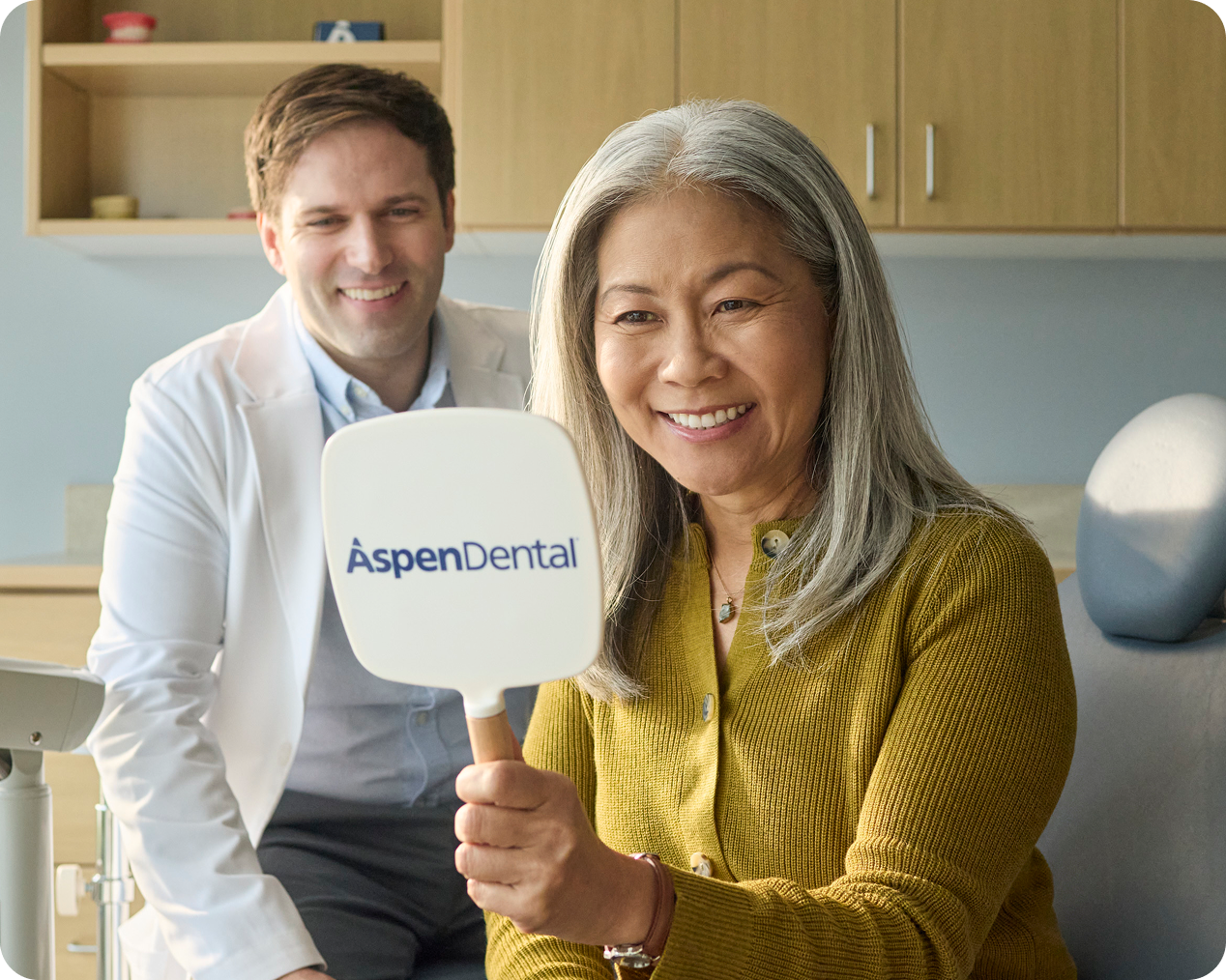Smiling older woman holding a Aspen Dental mirror and viewing her smile during a dental visit, with a dentist in the background.