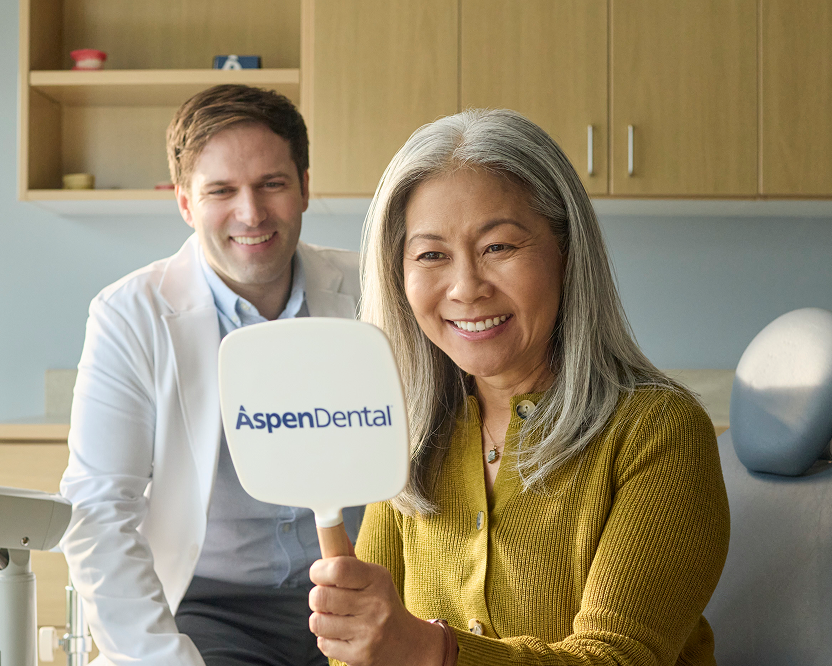 Alt text:
Smiling older woman holding an Aspen Dental mirror and viewing her smile, with a dentist in the background.