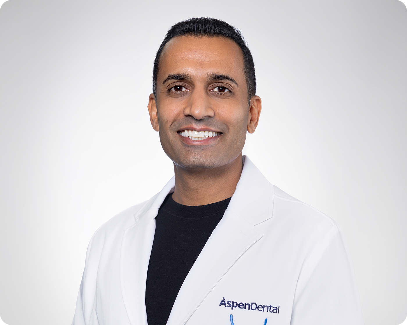 Portrait of a smiling Aspen Dental dentist wearing a white lab coat against a light gray background