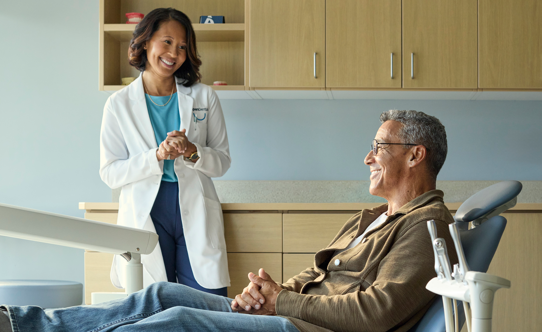 An Aspen Dental dentist standing beside a senior male patient reclining in a dental chair, smiling and discussing tooth extraction and cost details during a chairside consultation.