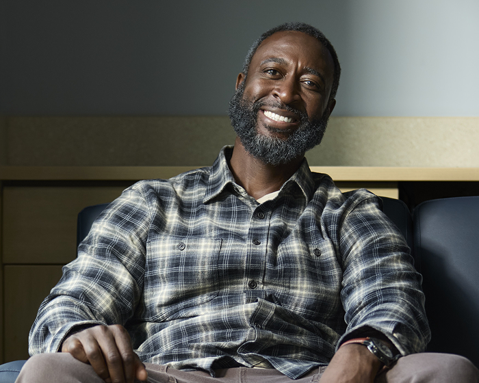 Smiling adult man seated indoors with a relaxed, confident expression.