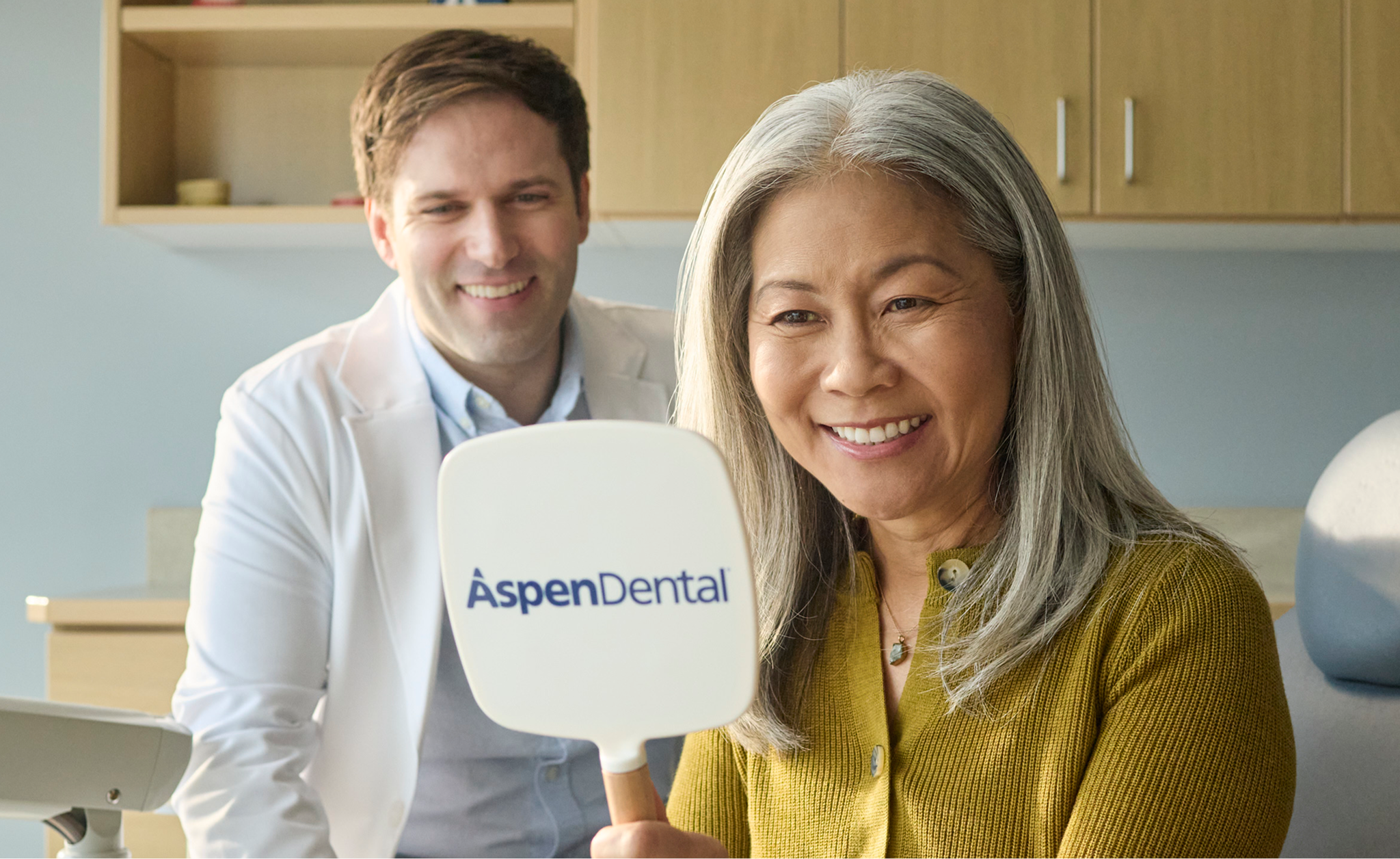 Smiling older woman holding an Aspen Dental mirror and viewing her smile during a dental visit, with a dentist in the background.