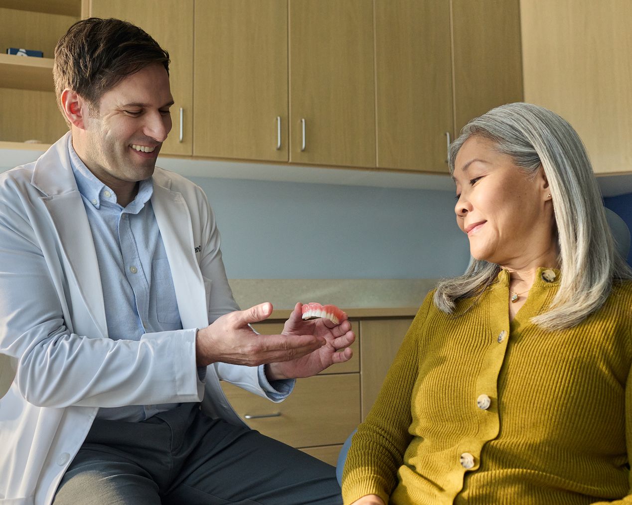 Alt Text (Recommended):
Aspen Dental dentist explaining denture options using a dental model during a consultation with a senior patient.