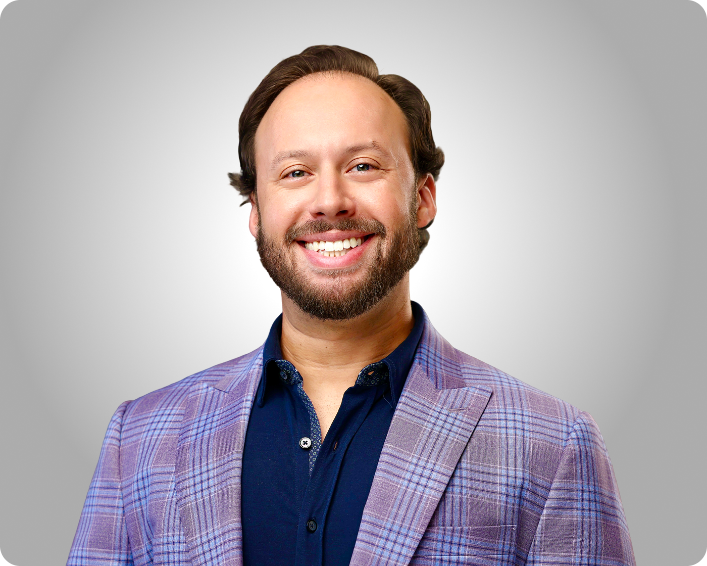 Professional headshot of a smiling man wearing a purple plaid blazer and navy shirt against a gray background