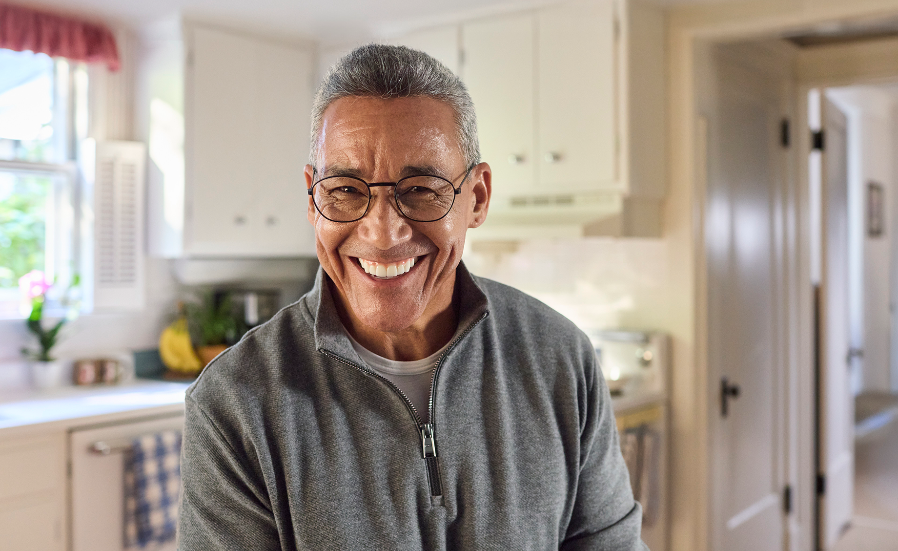 Alt text: A confident older man smiling brightly in his kitchen, showing off his new dentures after completing the adjustment process.