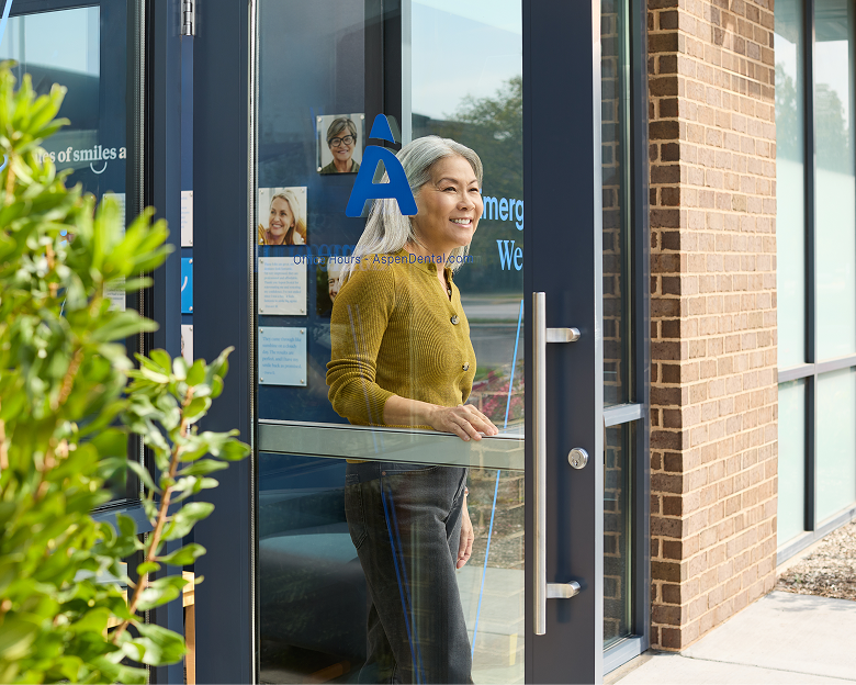 Smiling older woman leaving an Aspen Dental office through a glass door.