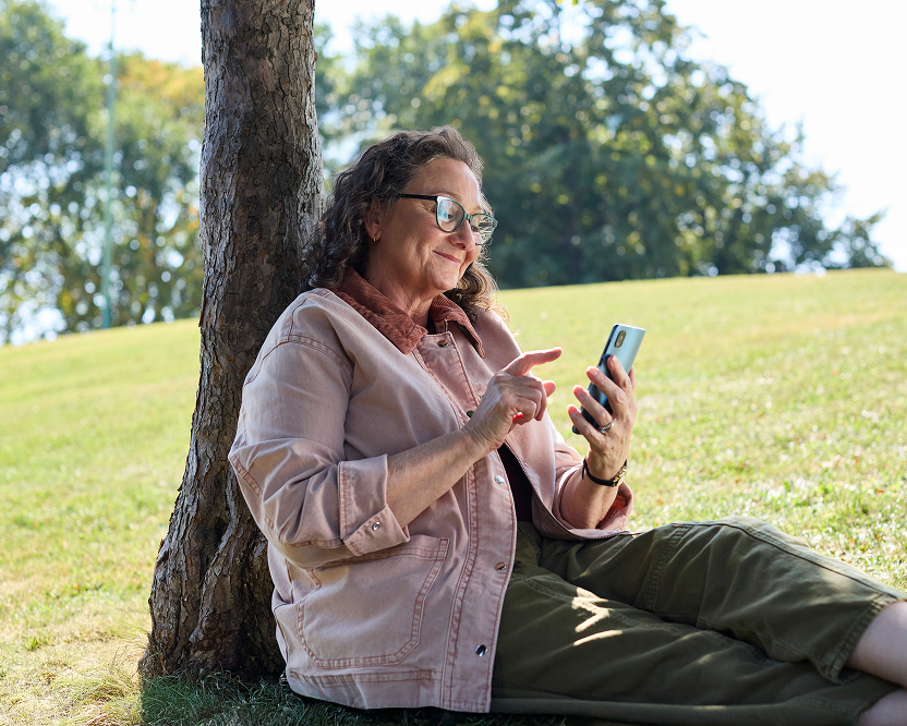 Alt text:
Smiling adult woman sitting outdoors under a tree while using a smartphone, representing managing dental care online.