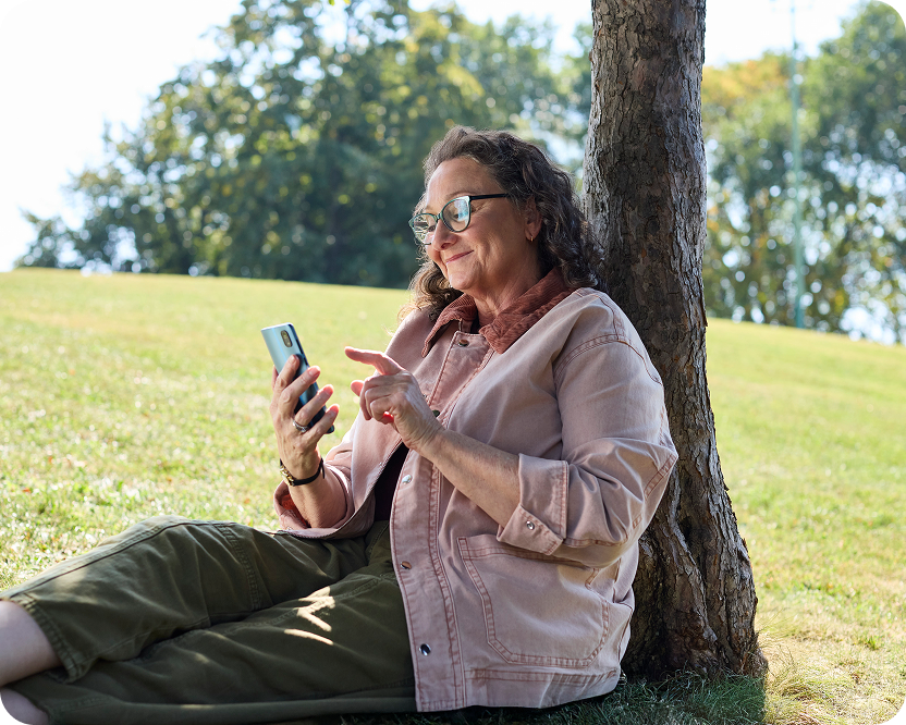 Alt text:
Woman sitting under a tree using a smartphone outdoors.