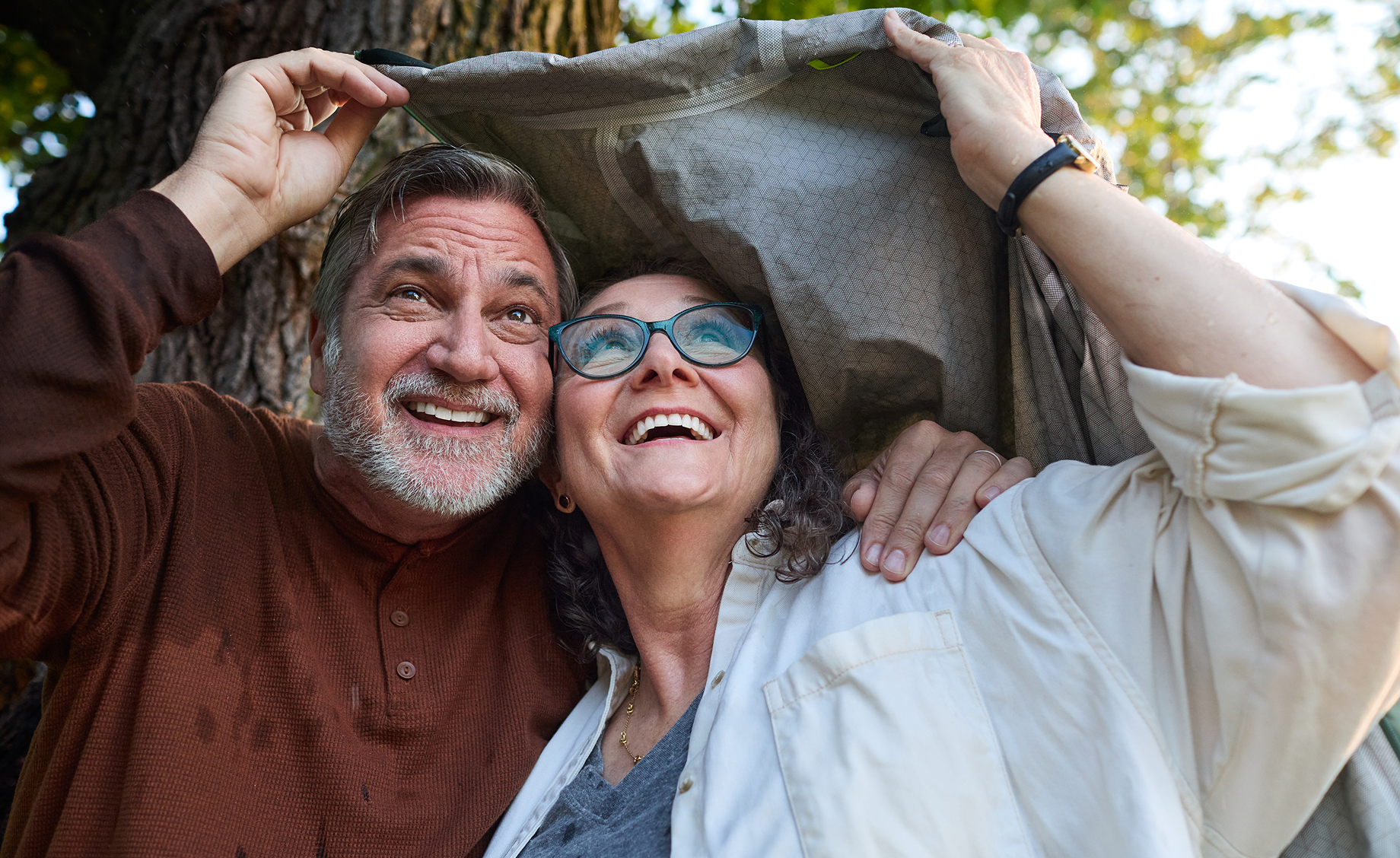 Smiling older couple holding a jacket overhead outdoors and looking up.
